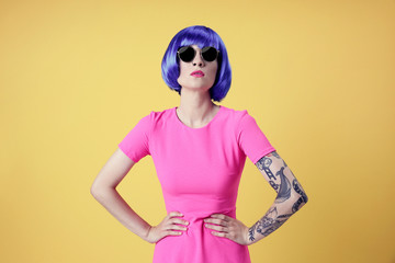 Beautiful young woman with tattoo wearing blue wig and sunglasses on yellow background