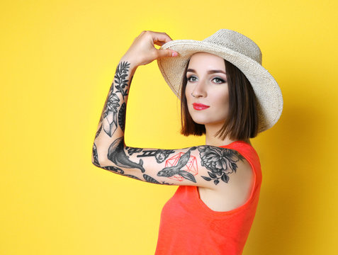 Beautiful young woman with tattoo wearing hat and posing on yellow background