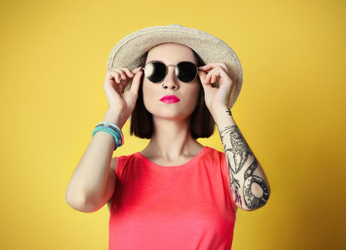 Beautiful young woman with tattoo and sunglasses wearing hat on yellow background