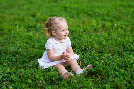 Baby girl in white dress sits and smiling on the fresh green grass