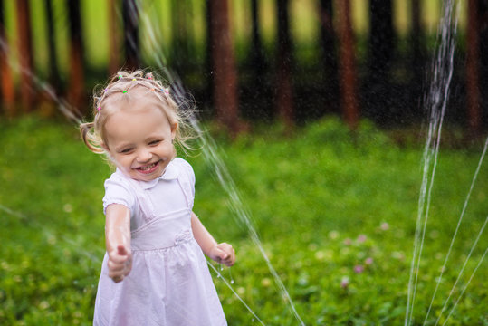 Little girl in white dress plays with sprinkler in the garden