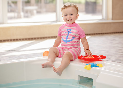 Portrait of white Caucasian baby girl laughing sitting on swimming-pool nosing looking in camera, training to swim, healthy active lifestyle
