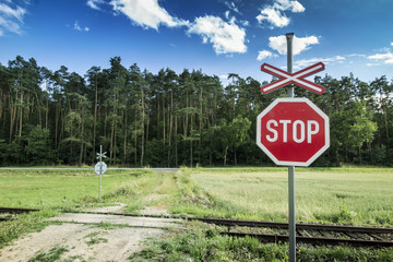 Stop Sign on Unguarded Rail Road. Road Across Rails with Field, Forest and Blue Sky.