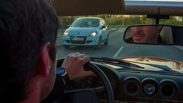 A man and woman are driving in an open roof car and we can observe a man in the rear mirror as he is saying something to his wife. Close-up shot. They are driving through town.
