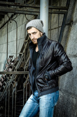 Obraz premium Bad boy concept. Portrait of brutal young man with short hair wearing black jacket and gray hat , Hipster style. Copy-space. Outdoor shot