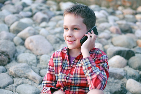 Caucasian Kid Talking On The Phone Sitting On The Rocks. Handsome Boy With Mobile Smartphone. Outdoors 