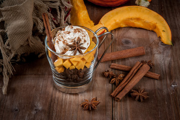 Concept: fragrant autumn drink - all you need for pumpkin coffee latte with whipped cream and spices - aniseed and cinnamon. On the wooden background, next to a warm blanket, spices and pumpkin.