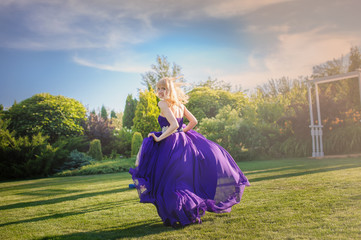 Blonde gorgeous woman in long purple luxurious dress posing in garden. Dress fluttering on wind.