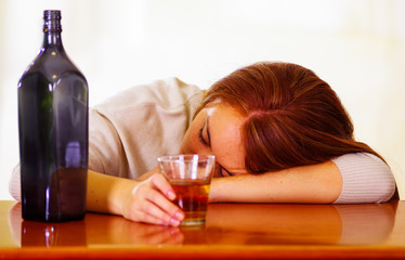 Attractive woman wearing white sweater sitting by bar counter lying over desk next to glass and bottle, drunk depressed facial expression, alcoholic concept