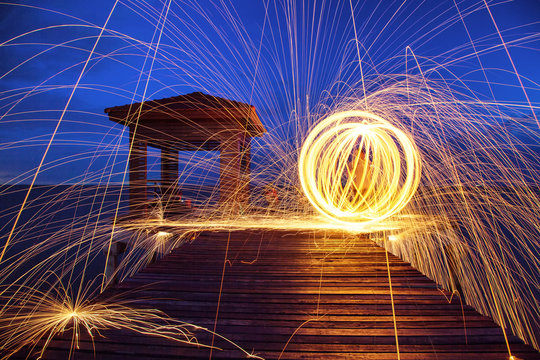 Hot Golden Sparks Flying from Man Spinning Burning Steel Wool on Wooden Bridge Extended into the Sea., Long Exposure Photography using Steel Wool Burning.