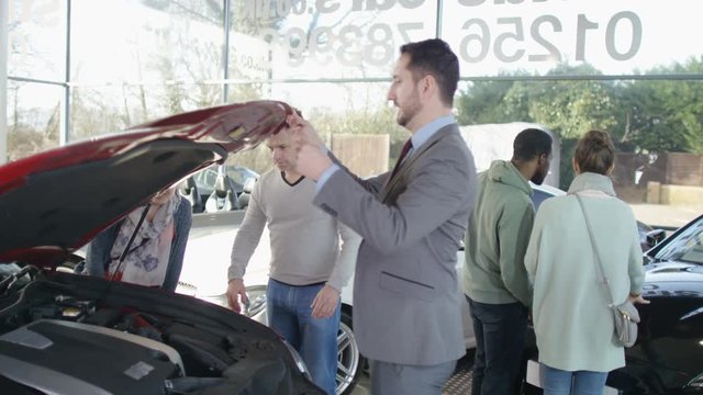  Salesman in car dealership with couple, showing them the engine of new car. 