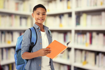 Cute boy with backpack holding book on blurred book shelves background. Library concept.