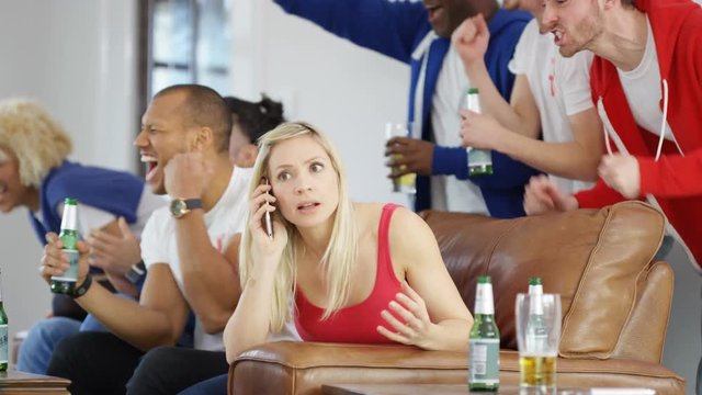  Woman talking on mobile phone while rowdy friends watch sports game on TV