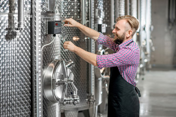 Handsome wine maker in working apron pouring wine to the glass at the wine manufacture with metal tanks for wine fermentation. Wine production at the modern factory