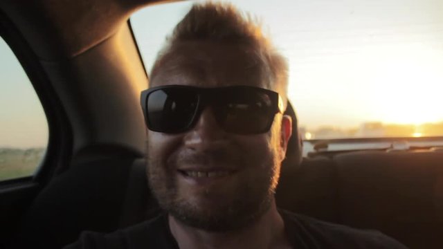 Young casual man with beard portrait sitting on the backseat of car. Urban male hipster in his 30s. Handsome young beared man smiling happy outside looking at camera. Sunset