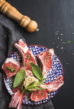 Raw uncooked lamb chops with herbs and spices on colorful plate over dark wooden background, top view, vertical composition