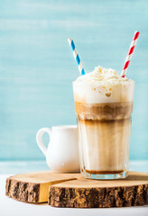 Latte macchiato with whipped cream in tall glass with two straws and pitcher on wooden board over...