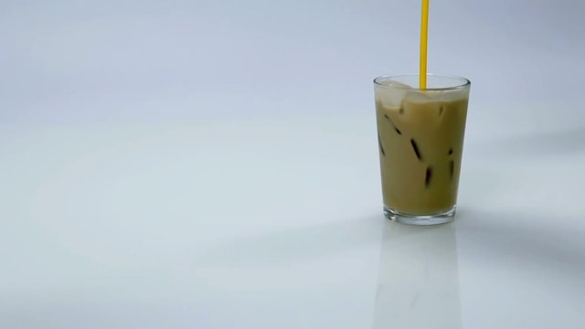 A glass with ice and cappuccino is standing on a table. Someone starts stirring the liquid in the glass with a yellow straw. Close-up shot
