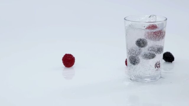 Raspberries and blueberries are falling into a cup of water which is standing on the table in the kitchen. Some water spills out. Close-up shot.
