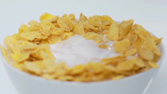 A very full cup with corn flakes and milk is standing on a table and some blueberries are falling in. Some milk and corn flakes fall out of the cup on a table. Close-up shot.
