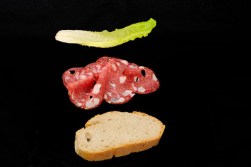 ingredienti per preparare un gustoso panino, fetta di pane, tre fette di salame spianata, foglia di lattuga, sfondo nero