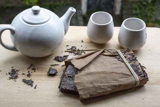 One Pack Of Black Chinese Tea With White Teakettle And Two Cups With Green Leaves As Background