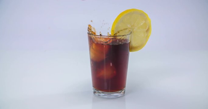 Someone throws a few ice cubes into a glass with some coke. A piece of lemon wheel is added on its rim.
