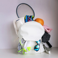 White leather bag (daypack) with clothing, sport shoes, battledore, grapefruit and sunglasses pocked out over the top