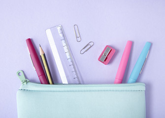 A pastel blue pencil case with stationery and pens spilling out on to a purple background