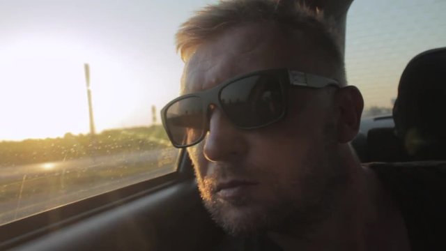 Young casual man with beard portrait sitting on the backseat of car. Urban male hipster in his 30s. Handsome young beared man looking at camera. Sunset