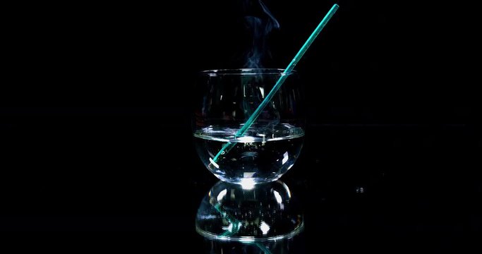 A glass which is half full with water is standing on a table in the kitchen. A green straw falls into it. Wide-angle shot.
