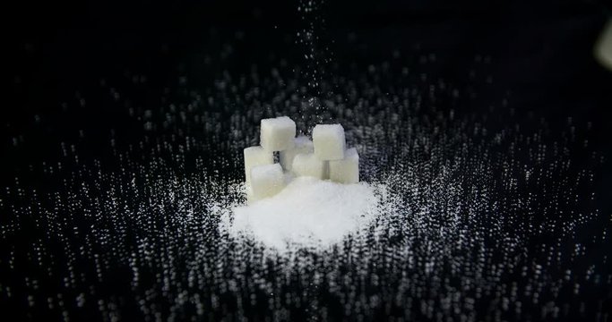 A few sugar cubes are lying on a table in the kitchen. Someone starts pouring grains of sugar on these cubes and past them. Close-up shot.
