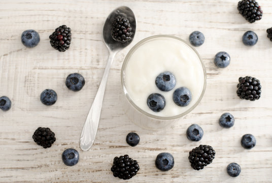 Top view of blueberries in yogurt, blackberries in a spoon and berries on the table, light wood background