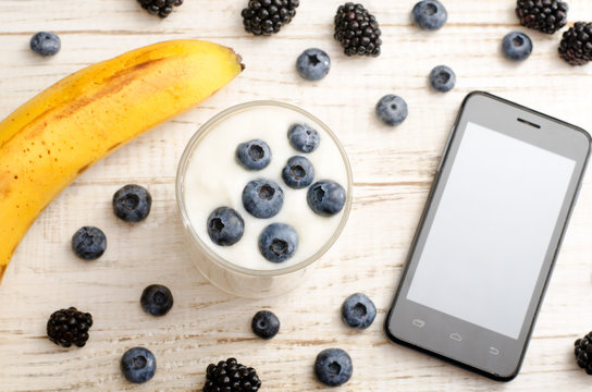 Yogurt with blueberries, ripe banana, smartphone and berries on the table