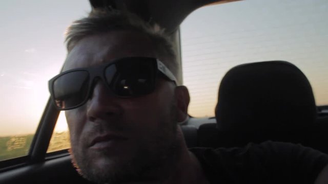 Young casual man with beard portrait sitting on the backseat of car. Urban male hipster in his 30s. Handsome young beared man looking at camera. Sunset