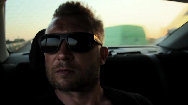 Young casual man with beard portrait sitting on the backseat of car. Urban male hipster in his 30s. Handsome young beared man looking at camera. Sunset