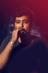 Young man smoking cigarette on dark background looking into came
