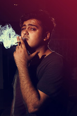 Young man smoking cigarette on dark background looking into came