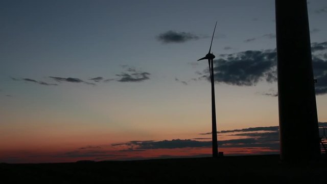 Widok na kręcące się śmigła wiatraka podczas zachodu słońca, pomarańczowe niebo i kilka chmur na niebie