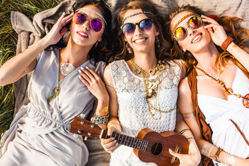 Three cute hippie girl lying on the plaid outdoors, best friends having fun and laughing, play ukulele, sunglasses, feathers in their hair, bracelets, flash tattoo, indie, Bohemia, boho style top view - obrazy, fototapety, plakaty