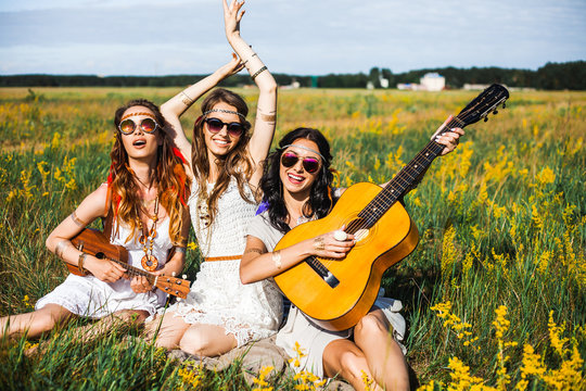 Three cute hippie girl sitting on grass outdoors, best friends having fun and laughing, playing the guitar and ukulele, feathers in their hair, bracelets, flash tattoo, indie, Bohemia, boho style