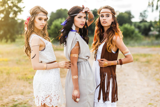 Three cute beautiful hippie girl walking along the road, outdoors, best friends are traveling, make-up, long hair, feathers in their hair, bracelets, flash tattoo, indie, Bohemia, boho style