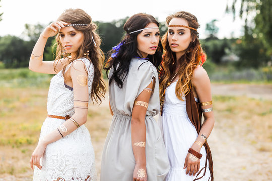 Three cute beautiful hippie girl walking along the road, outdoors, best friends are traveling, make-up, long hair, feathers in their hair, bracelets, flash tattoo, indie, Bohemia, boho style