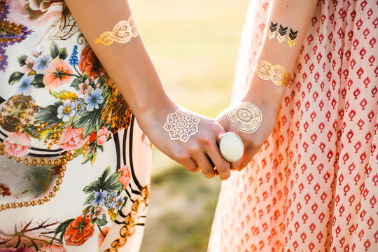 Two cute hippie girl in the setting sun, outdoors, holding hands, best friends, feathers in long hair, bracelets, flash tattoo, indie, Bohemia, boho style dresses, trendy accessories