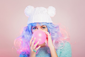 Cute young woman with pastel hair and knitted beanie hat blowing a pink balloon
