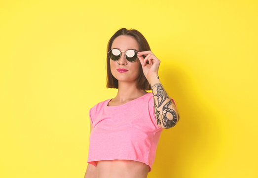 Beautiful young woman with tattoo wearing sunglasses and posing on yellow background