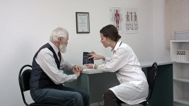 Woman doctor talking with senior patient in the office and showing him tablet with mri.