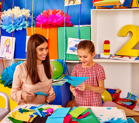 Child girl with teacher are making something out of colored paper on table in primary school....