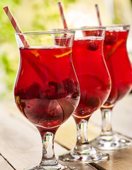 On wooden boards are group of ice cold beverage glasses with red raspberry lemon cocktail . Alcohol cocktail beverage hundred two hundred ninety nine. Cocktail outdoor in garden. Glasses stand in row.