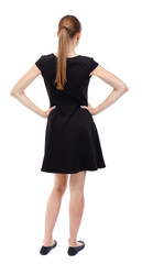 Fototapeta premium back view of standing young beautiful woman. girl watching. Rear view people collection. backside view of person. Blonde in a short black dress standing with hands on waist. 